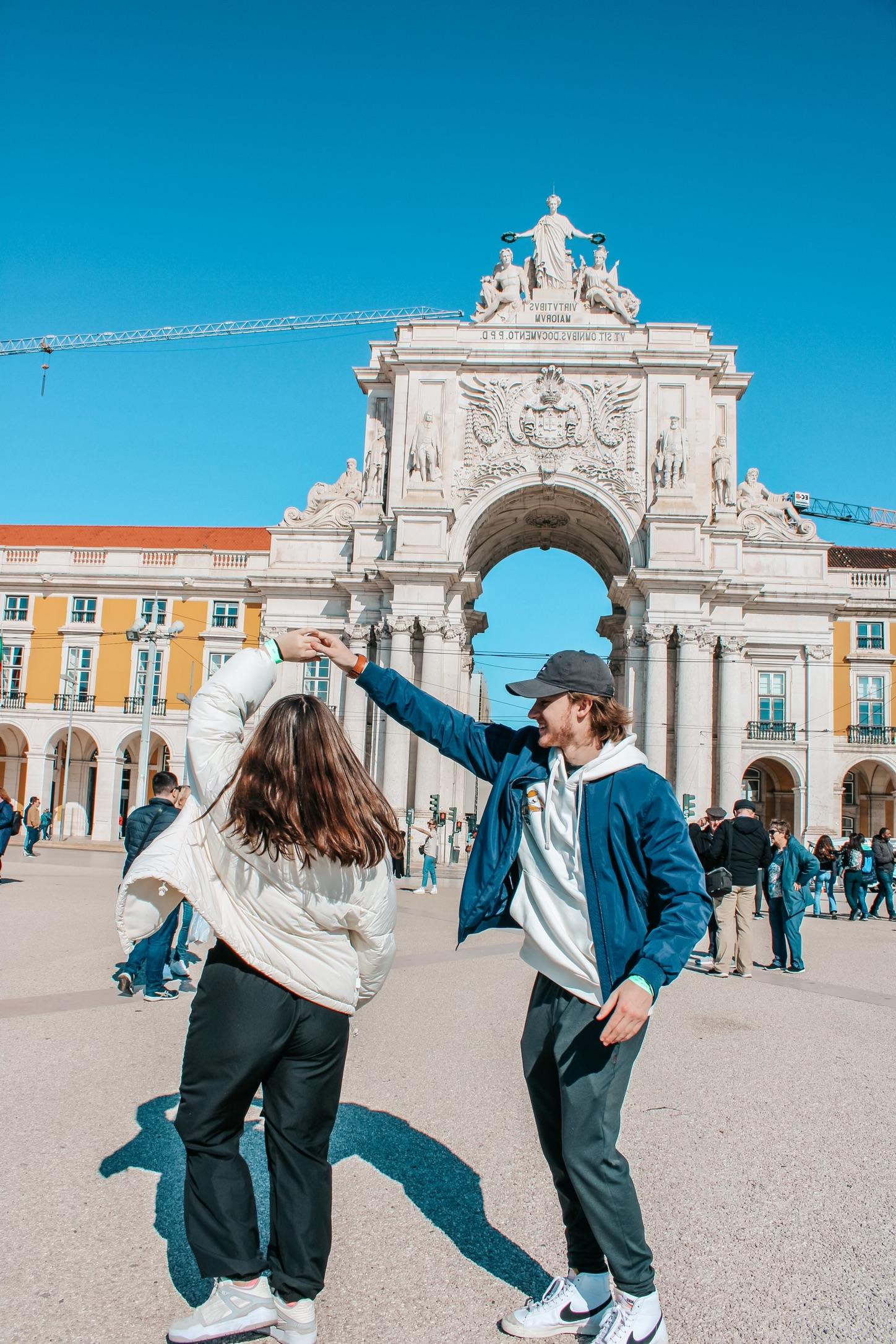 在Arco da Rua Agusta前，穿着白色夹克的女子与戴着黑色棒球帽和蓝色夹克的男子旋转